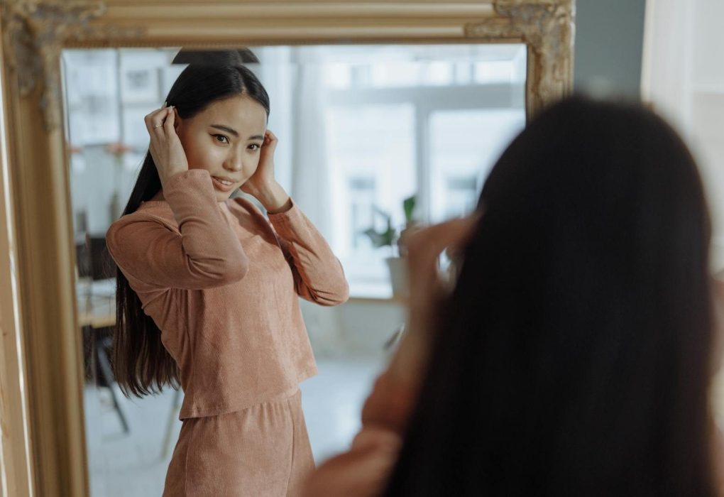 Elegant young woman in brown dress adjusting her hair while looking at her reflection in a mirror indoors.