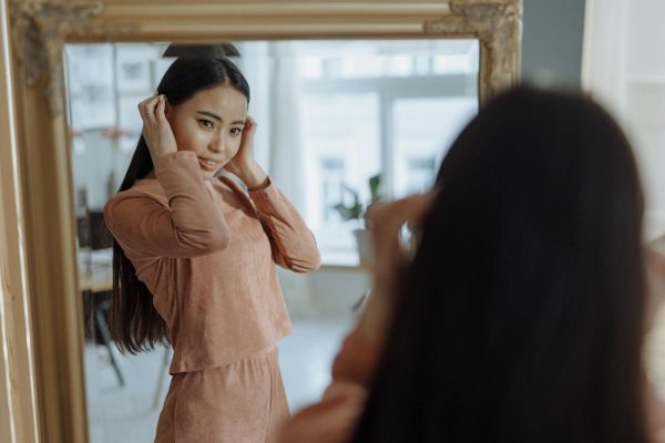 Elegant young woman in brown dress adjusting her hair while looking at her reflection in a mirror indoors.